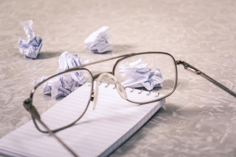 Pair of glasses sitting on top of a pad of paper with balls of paper in the background.