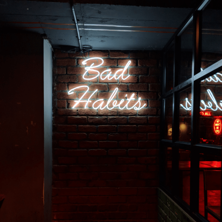 White neon sign that says "Bad Habits" in a script font against a dark background.