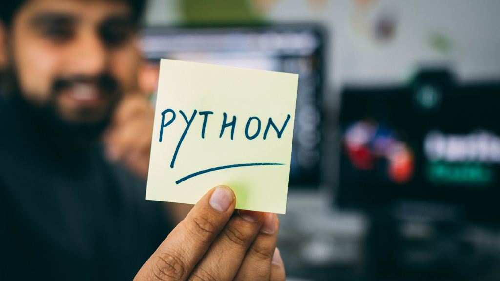 Python lead job description | Picture of a post-it note with the word “Python” on it, against a blurry background of a man with a bear, who is holding the post-it.