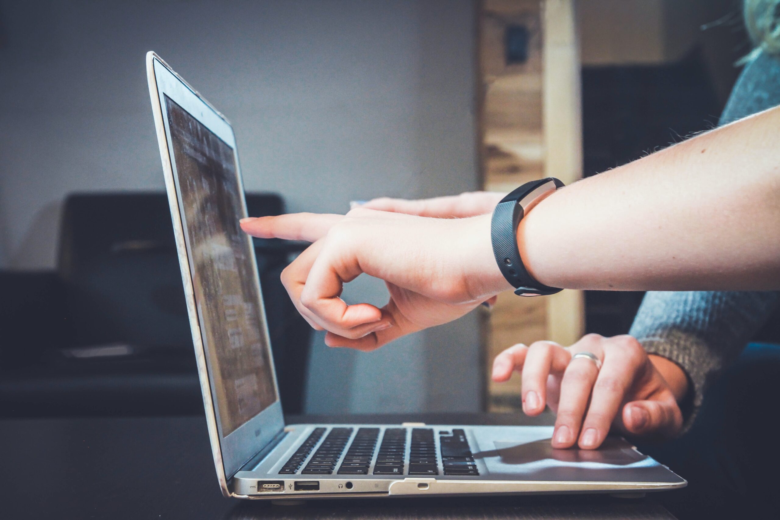 How to hire TypeScript developers | Picture of someone's left hand pointing at a laptop screen, with another person in the background using the touch pad on the same laptop.