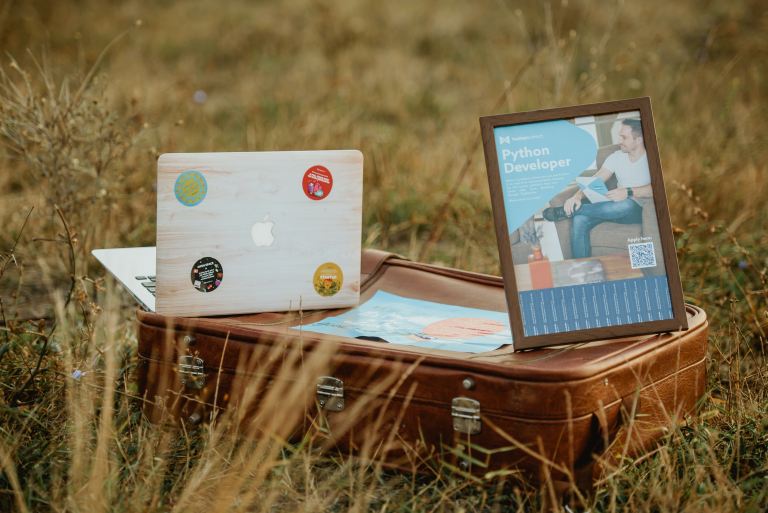 Hire dedicated Python developers | Image of a computer and a frame with a picture that says ‘Python Developer’, sitting on top of a leather case in the grass