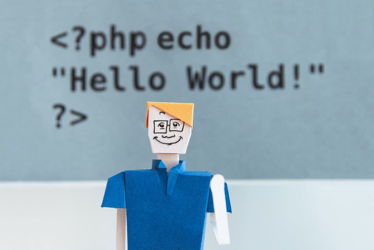 PHP developer job description | Folded paper man with yellow hair and a blue shirt against a blurred background of PHP code.