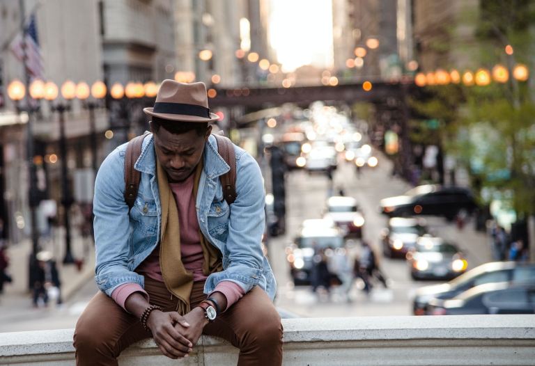 Introverted engineering manager | Man sitting on a bridge above a city street, wearing a hat, blue button-down shirt, yellow tee, and brown pants, clasping his hands and looking down at the ground.