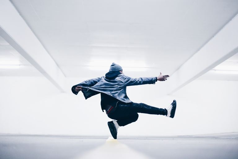 Person in a beanie, denim jacket, black pants, and two-tone shoes jumping up in the air in a white room.