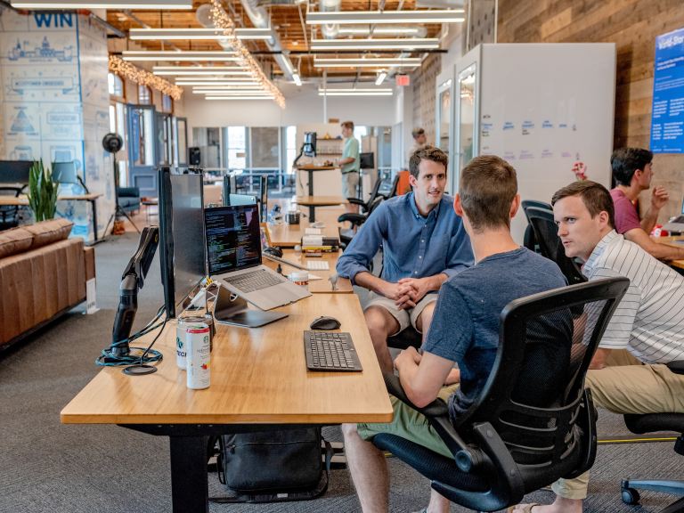 App dev agency | Picture of a bright, vibrant office with three men in the foreground who are sitting around a desk.
