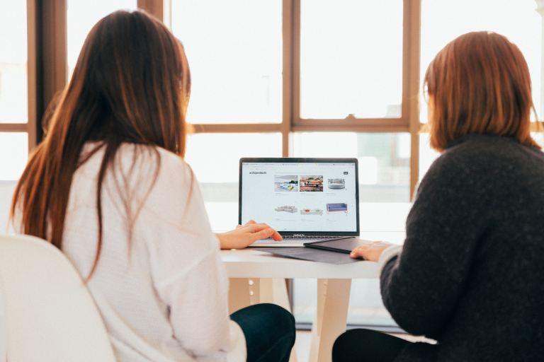 Hire web development company | Image shot from behind of two women sitting at a table and looking through designs on a computer.