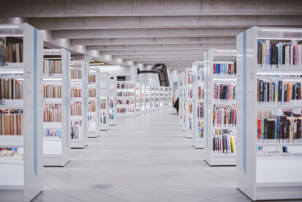 Custom component libraries vs off-the-shelf solutions | Wide aisle in a library with white shelves to either side filled with books; cement beams across the ceiling; and bright light.