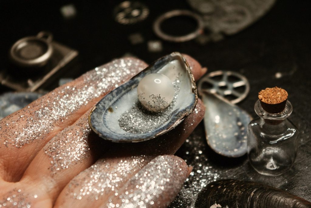 Perl developer job description | In the foreground, a hand covered in silver glitter holds an open shell with a pearl. In the background, there are small corked glass bottles, gears, and assorted trinkets.