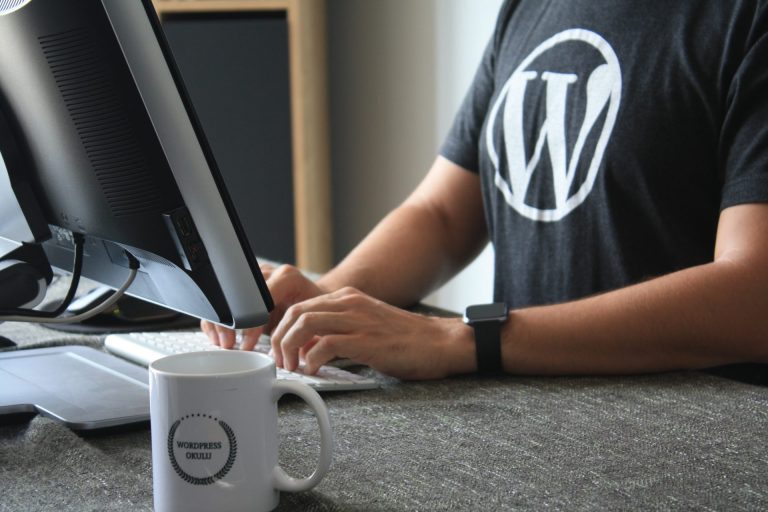 Hire dedicated WordPress developers | Torso shot of a person sitting at a desk with a desktop computer, wearing a dark grey shirt with the WordPress logo on it.