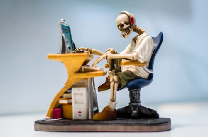 Skeleton in office attire sits at a desk with a laptop