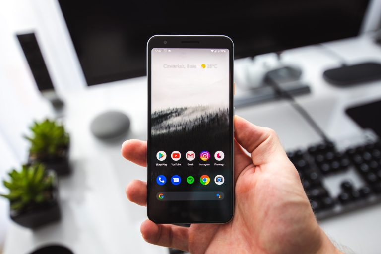 Android Engineer Job Description | Person holding an Android phone in their right hand with a blurred desk setup in the background.