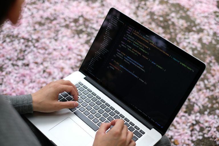 Next js interview questions | Person wearing a grey shirt with a silver laptop balanced on their knees. On the screen is code, and in the background is the ground covered in flower petals.