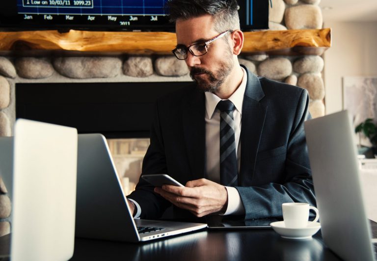 Photo of a man in a suit, sitting at a desk in front of a laptop with a phone in his hand.